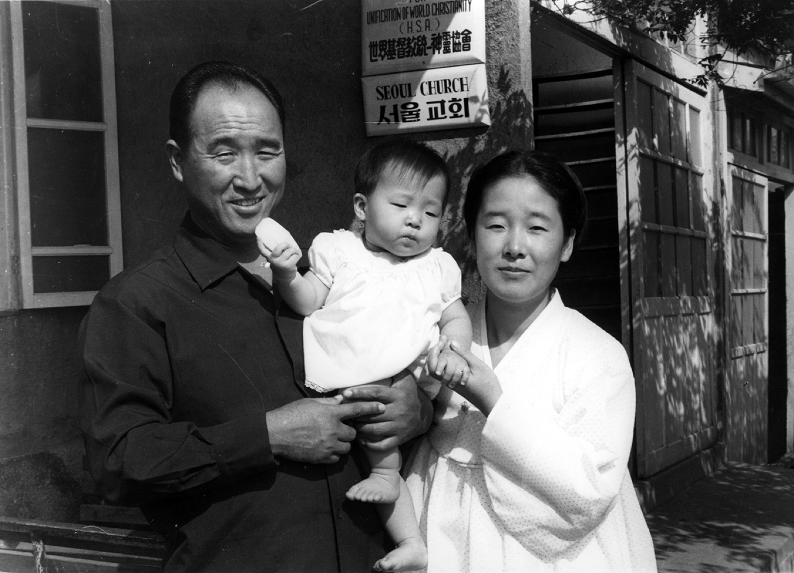 True Father holding a baby beside True Mother, black-and-white archive photo, 1965